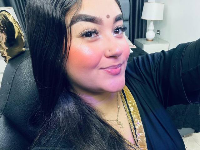 AnaShalimarr mit dunklem Haar, rosiger Wangen, schwarzen Augenbrauen, Nasenpiercing, goldener Halskette, schwarzem Oberteil, lächelnd, hält ein Telefon, macht ein Selfie, ein Stuhl ist sichtbar, eine weiße Lampe im Hintergrund..