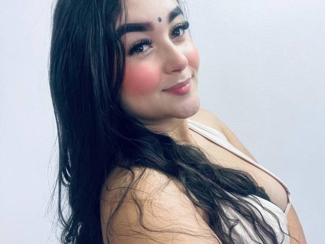 AnaShalimarr has long black hair, pink blush on her cheeks, and a small beauty mark above her right eyebrow. She's wearing a white top, showing her boobs. Smiling.