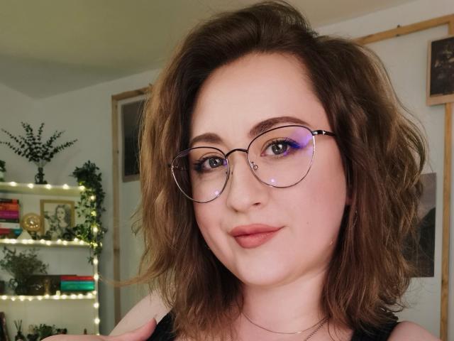 LuxNoir has wavy brown hair, purple eye makeup, and wears glasses. She's smiling with pink lips, wearing a black top, showing her boobs in a selfie.