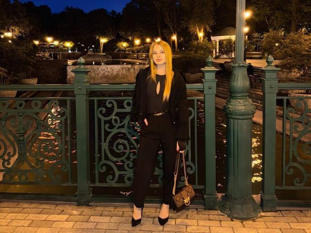 AngyyEyess stands in front of an ornate green fence, wearing a black outfit with a belt, black heels, and holding a patterned handbag, with long blonde hair.