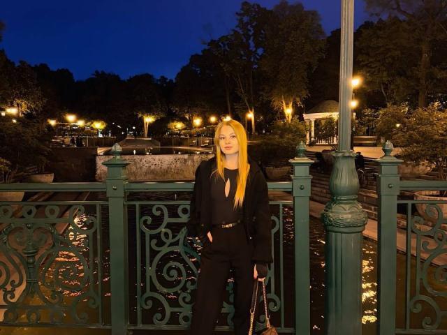 AngyyEyess with long blonde hair, black outfit, and hands in pockets, stands on a green ornate bridge at night, holding a black purse, under streetlights.
