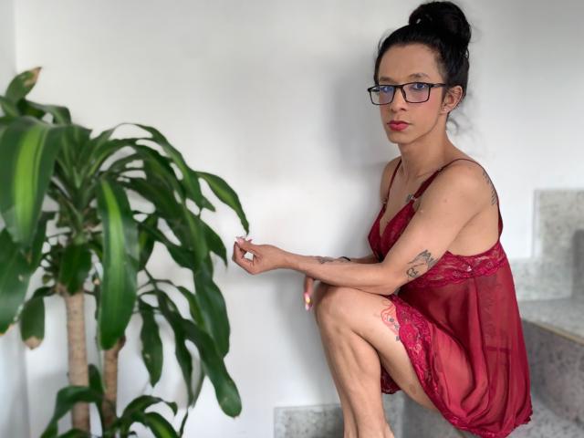 Daslyroze69, wearing red lace lingerie, sits on stairs with one knee up, touching a green plant, has tattoos, glasses, and black hair in a bun, looking at camera.