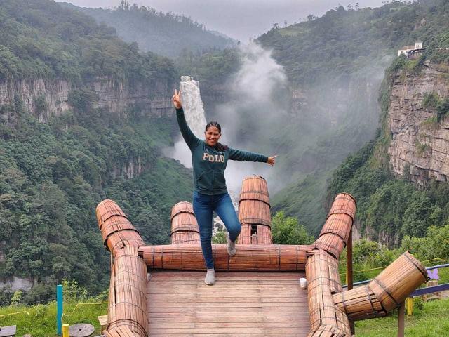 PauletteLoveX, with a big ass and a happy smile, jumps on a bamboo bridge, holding up a white rock, wearing a green "FUJI" sweater and blue jeans.