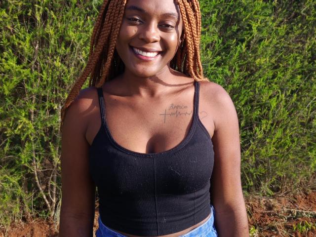 FireGal com cabelo trançado marrom, sorrindo, vestindo uma regata preta que mostra seios pequenos, tatuagem no peito, jeans azuis, em pé ao ar livre, fundo verde e cheio de folhagem, ensolarado, expressão feliz..