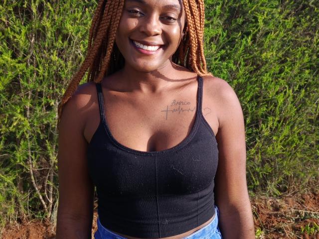 FireGal, com pele escura, sorrindo, vestindo uma camiseta preta que mostra seus seios e jeans azuis, tem cabelo trançado laranja, uma tatuagem no lado direito do peito e está em um ambiente externo..