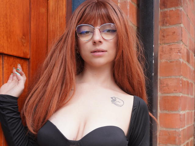 Anntonella Watson, con cabello rojo largo, gafas, un top negro sin tirantes que muestra un busto grande, un tatuaje en el pecho derecho, de pie junto a una pared de ladrillos, con una puerta de madera a la izquierda..