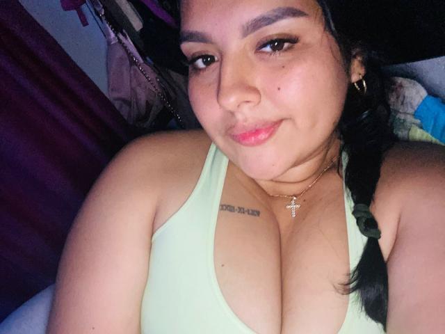 TindraZavaleta heeft een medium bruine huid, zwart gevlochten haar, een groen tanktopje en een kleine kruisbedel. Ze draagt een tattoo boven haar decolleté en haar grote borsten zijn zichtbaar. Ze glimlacht..