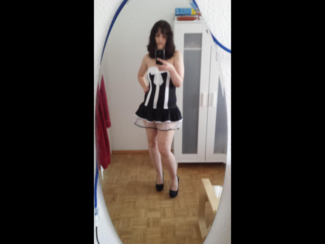 Mirror selfie of sweetSilvana-hot in a black-and-white maid dress with frilled skirt, black heels, and wavy brown hair, standing in a room with wooden floor.