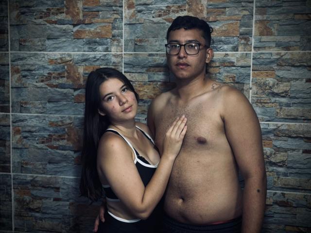 BellaThorSexUnlimit with small tits, black tank top, and long brown hair stands next to shirtless BellaThorSexUnlimit with glasses, dark hair, and chest hair, touching chest.