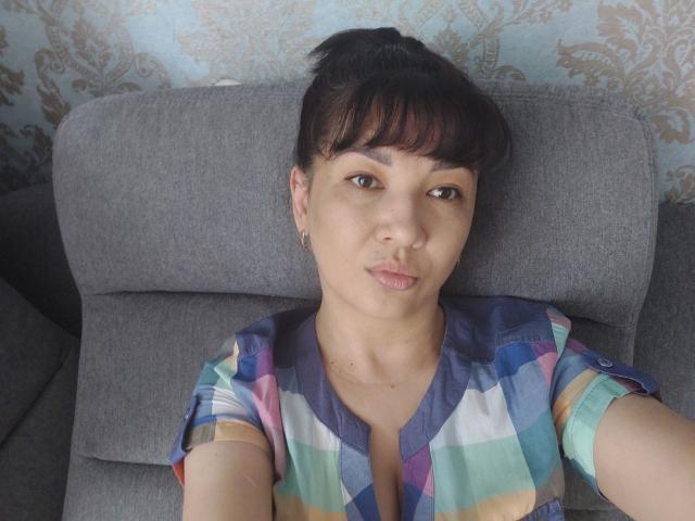 SelenaFly with dark hair in a ponytail, wearing a colorful checkered shirt, sitting on a gray couch, pouting her lips, medium-sized tits, light brown skin, and brown eyes.
