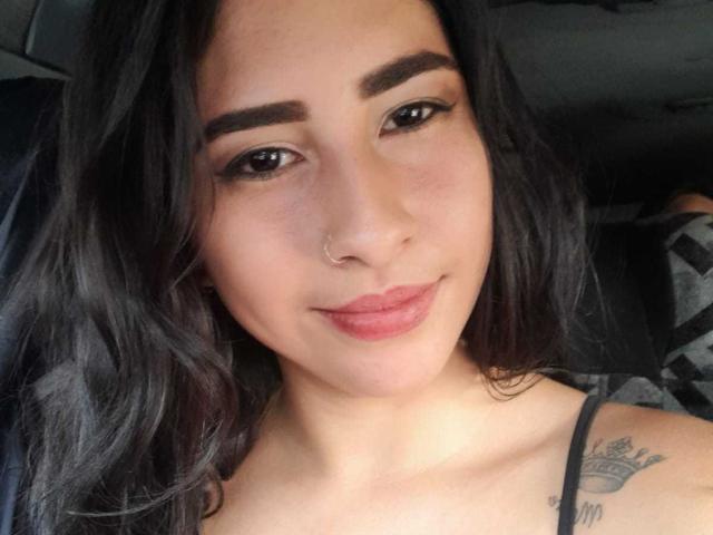 Katnis has dark wavy hair, thick eyebrows, a nose ring, and a tattoo on her right shoulder. She's smiling with pink lipstick and wearing a black spaghetti strap.