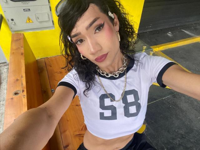 SweetSamyPrincipal with curly black hair, wearing a white sports jersey with number 58, chain necklace, and dark makeup, taking a selfie in a yellow and wood setting.