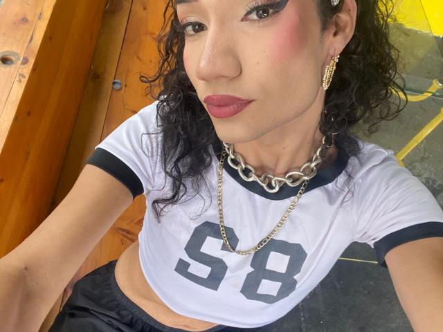 SweetSamyPrincipal with curly black hair, dark makeup, pink blush, red lipstick, wearing a white sports shirt with "58" and chain necklace, showing off gold earrings and a confident smirk.