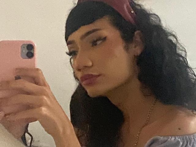 SweetSamyPrincipal with curly black hair, pink headband, and off-shoulder top, taking a selfie with a pink phone, showing off her pouty ass-kissing lips.