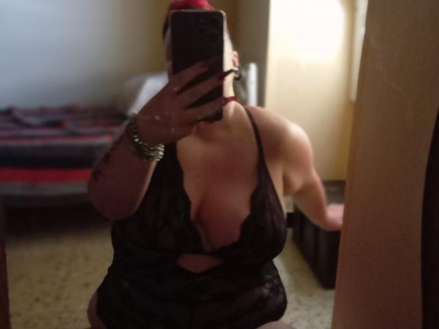 MmeCauchemar con cabello rojo, busto grande, usando lencería de encaje negro, sosteniendo un teléfono, selfie frente al espejo, pulsera en la muñeca, fondo borroso, de pie en un dormitorio, tomando una foto..