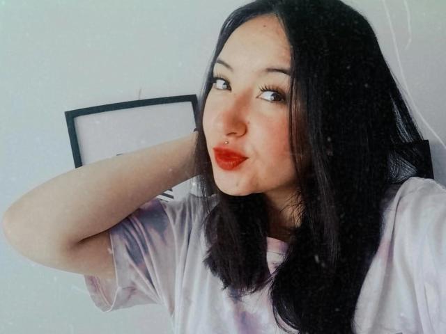 SusanMonroy with dark black hair, red lipstick, nose ring, and pale skin, wearing a pinkish shirt, posing with one arm behind her head. Her tits are partially visible.