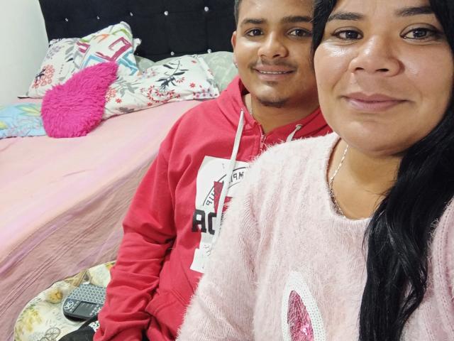 LatinStyleX with medium brown skin, black hair, and pink fuzzy top, sitting next to LatinStyleX with medium brown skin, black hair, and red hoodie, on a pink bed.