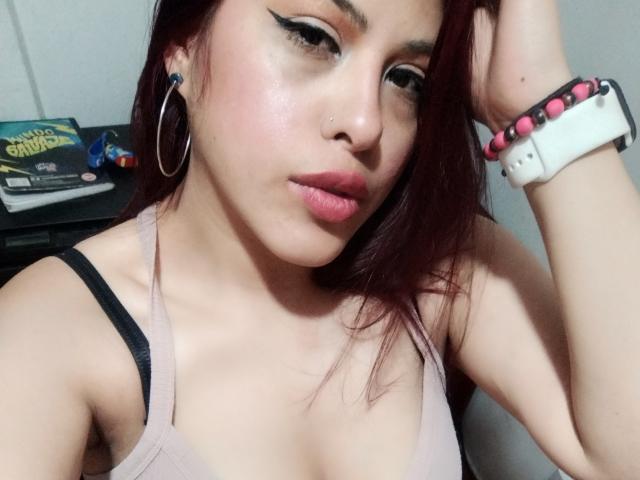ElaiaaBakerr with red hair, hoop earrings, black eyeliner, nose ring, pink lipstick, light skin, pink wristband, beige tank top, showing her chest and looking at camera.