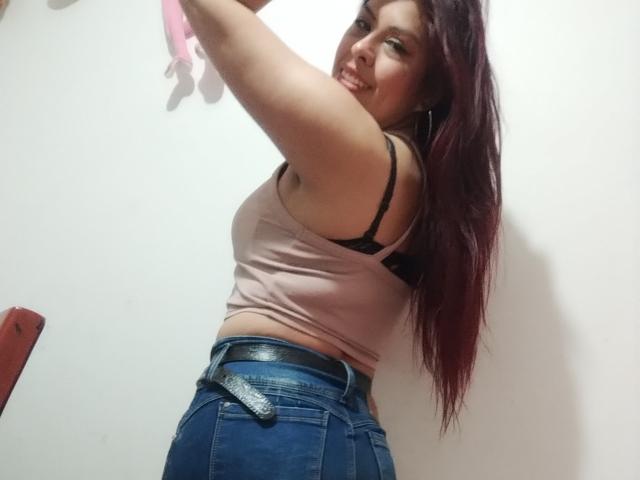 ElaiaaBakerr with red hair, smiling, wearing a pink tank top showing black bra, and blue jeans with a black belt, raising her arms. Her tits are visible.