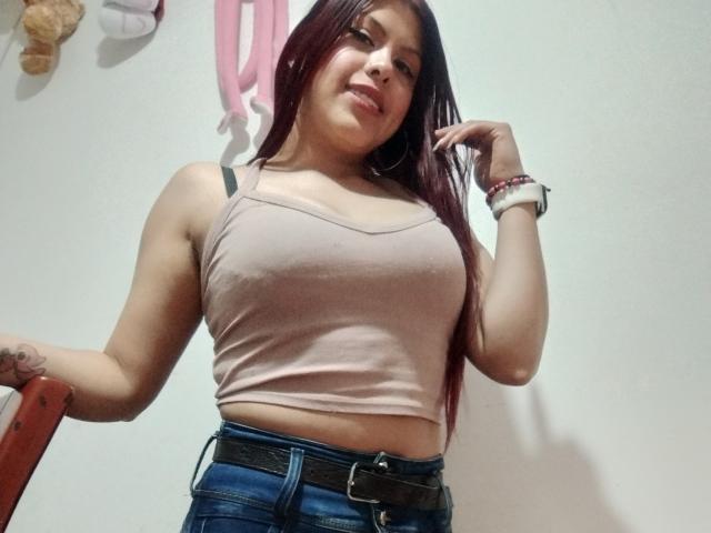 ElaiaaBakerr with long red hair, wearing a pink tank top showing her tits, and blue jeans with a black belt, smiling and holding her hair.