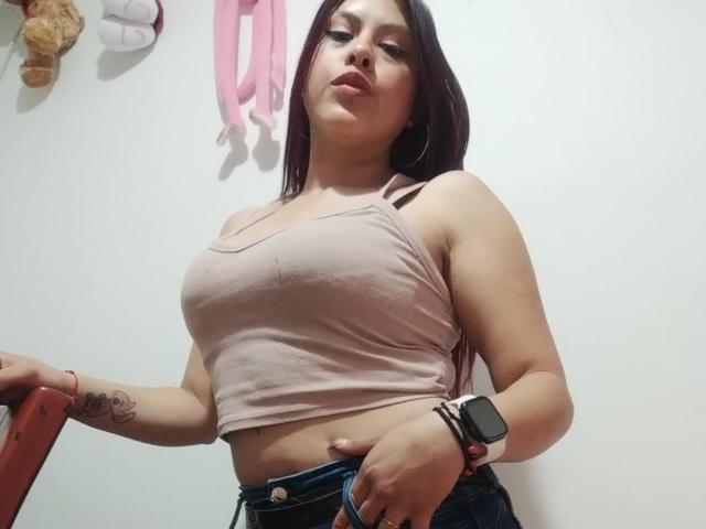 ElaiaaBakerr with large tits in a pink tank top, blue jeans, and a smartwatch, standing, looking down, long dark hair, tattoo on right arm, white wall background.