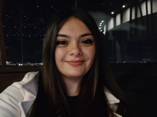 KatherinLatin has long brown hair, fair skin, and a slight smile. She's wearing a white jacket with black shirt, and has small, cute tits. Night city lights in background.