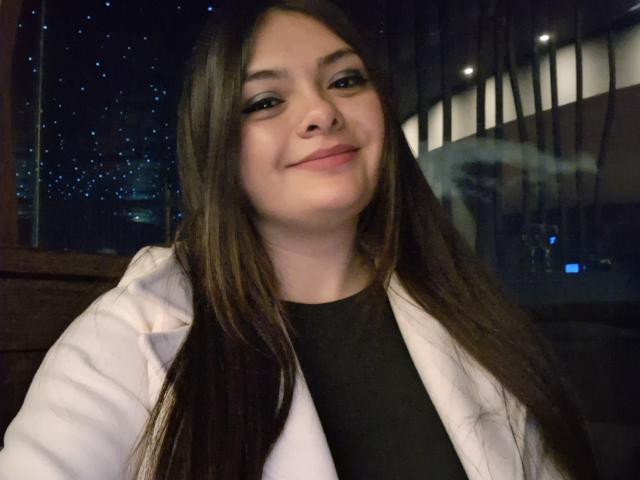KatherinLatin has long brown hair, a cute smile, and is wearing a white jacket over a black shirt. She's looking at the camera with a slightly upturned nose.