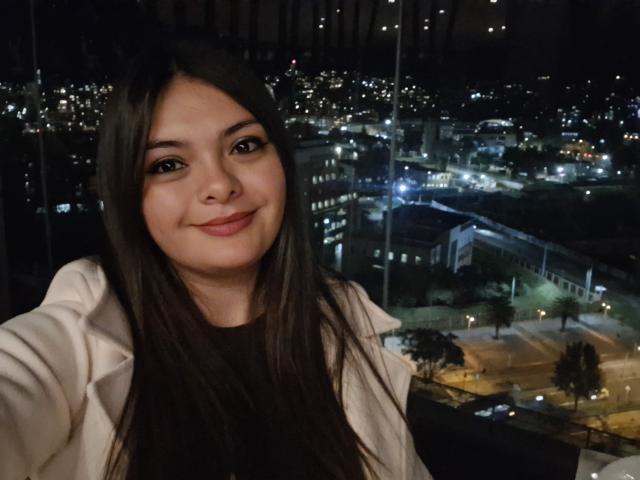 KatherinLatin has long dark hair, a cute face with a small smile, and is wearing a white jacket. She's taking a selfie at night with city lights in the background.