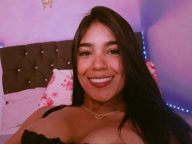 EmilyVio with long black hair, smiling, wearing a black lace bra, showing her large tits, a gold necklace, and pink floral pillows in the background.