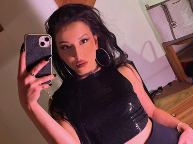 MissKataleya-hot with black hair, wearing a shiny black crop top, holding a phone, large hoop earrings, red lipstick, and a ring, taking a selfie in a mirror.