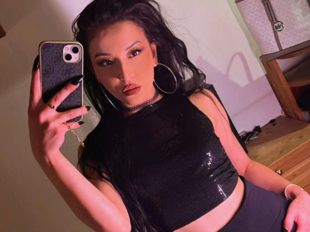MissKataleya-hot with long black hair, wearing a shiny black crop top, large hoop earrings, and holding a phone, showing off her tits in a mirror selfie.