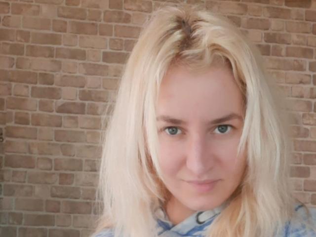 Xenya69 has blonde hair, green eyes, and a subtle smile. She's wearing a blue and white patterned shirt. Her ass is not visible. She's a cute, sexy chick.