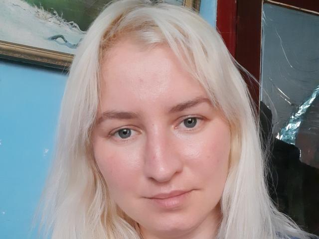 Xenya69 has pale skin, blue eyes, and long, straight, white-blonde hair. Her face is slightly flushed, with a neutral expression. Her tits are not visible.
