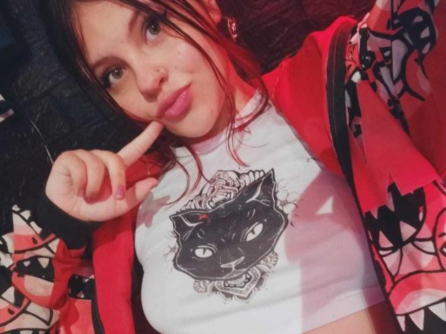 Sophie Hill, con una giacca a motivi rossi e neri, una t-shirt bianca con una grafica di un gatto nero, un dito sulle labbra, rossetto rosa, capelli castani, che guarda la fotocamera, seno di medie dimensioni, carina..