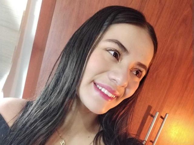 OshumBae has long black hair, brown eyes, and a cute smile with pink lipstick. She's wearing a black top and a gold necklace. Her tits are small and she's cute.