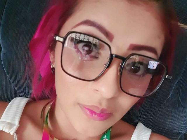 LizBonnet with pink hair, wearing glasses, reflecting a selfie in her eyes, pink lips, white straps, and a green necklace. Her reflection shows her looking at the camera.
