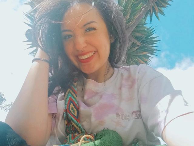 SusanMonroy with a nose ring, smiling, wearing a tie-dye shirt, holding colorful fabric, sunlight on her dark hair, blue sky in background, looking happy and relaxed.