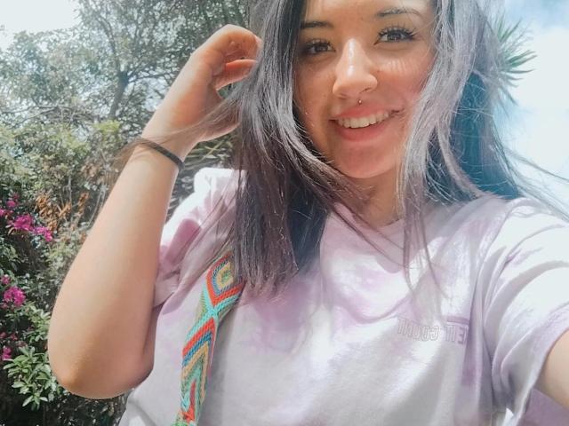 SusanMonroy with long brown hair, smiling, nose ring, wearing a tie-dye shirt, right hand in hair, purple and green patterns, sunlight, pink flowers in the background.