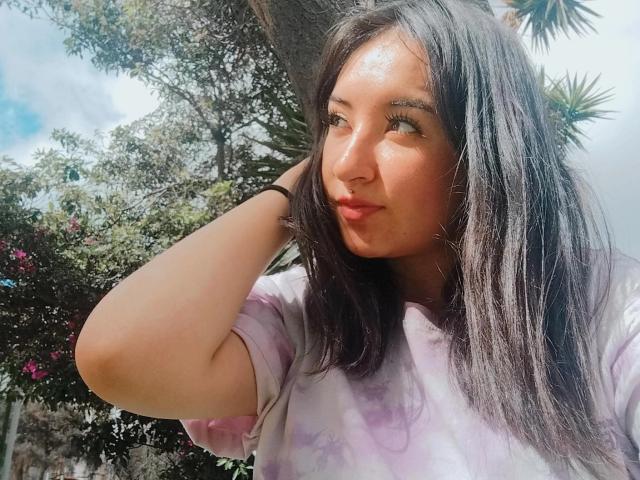 SusanMonroy with shiny brown skin, black straight hair, and a purple tie-dye shirt, slightly pouting with her hand in her hair, standing outdoors under a tree.