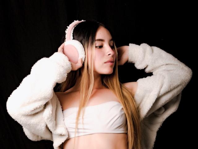 CandySays with long blonde hair, wearing pink fluffy headphones, white tube top, and fluffy white jacket, hands on head, looking down, black background.