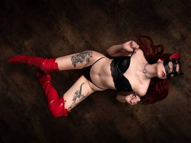 CherrySmarry-hot with red boots, black latex bra and thong, cat mask, and tattoos, lying on dark floor, showing her tits and ass, with long red hair.