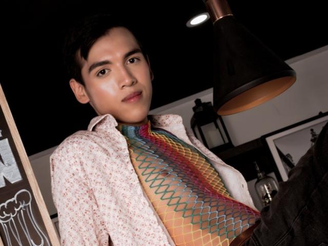 TheoBlake with short black hair, wearing a floral shirt and colorful, patterned tie, under a black lamp. He has a cute, slightly smiling face with medium skin.
