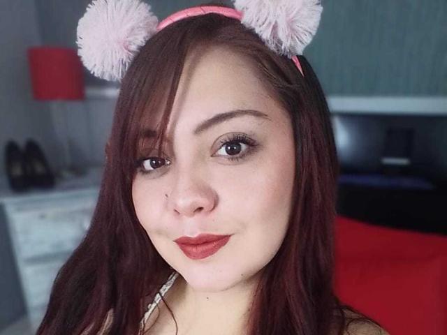 AmatisthaBlend with pink fluffy cat ears, red lipstick, and brown eyes, smiling slightly, brown hair, pale skin, small nose, wearing a pink headband, in a bedroom.