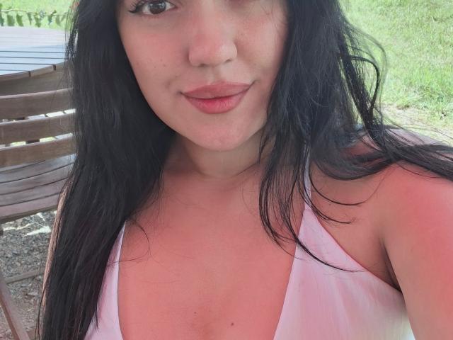 LauraRichis has long black hair, pink lips, and a slight smile. She's wearing a white halter top, showing her small tits and pink nipples. Outdoor selfie.