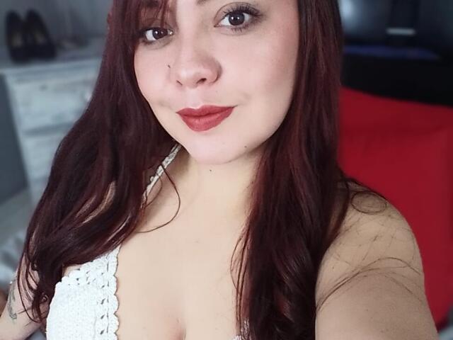 AmatisthaBlend with red hair, fair skin, and red lipstick, wearing a white lace top, showing her tits in a selfie. She has a subtle smile and dark eyes.