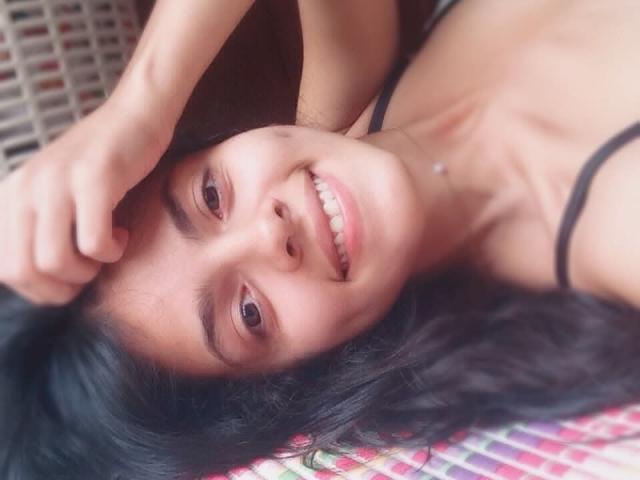 NanyTonelly's smiling face with black hair, brown eyes, and small nose, lying down with a hand in her hair, showing teeth and wearing a black strap.