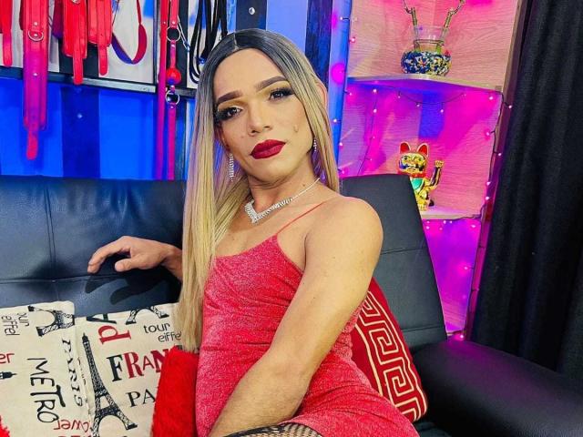 VeruzkaRose with long blonde hair, red lipstick, and a sparkly red dress, sitting on a black leather couch, holding a red pillow, wearing a diamond necklace.