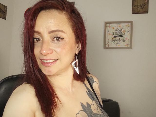 CatalinaMonyque has red hair, a tattoo on her shoulder, and is smiling with pink lipstick. She's wearing white, diamond-shaped earrings and a black strap top.