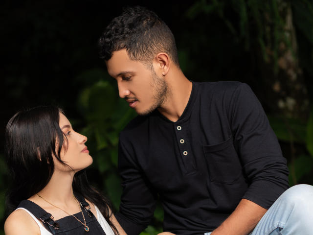 EimyAndMaxi with dark hair and white top, eyes closed, sitting. EimyAndMaxi with short curly hair, black shirt, looking down, hand on her shoulder. Forest background.