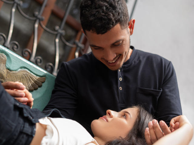 EimyAndMaxi with short curly brown hair, black shirt, smiling, holding head of EimyAndMaxi with straight dark hair, white top, lying down, looking up at each other.